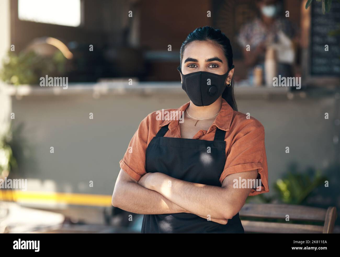Covid-Regeln folgend. Eine junge Frau, die mit gefalteten Armen vor ihrem Restaurant steht und eine Gesichtsmaske trägt. Stockfoto