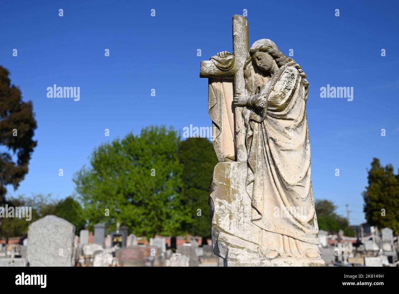 Alte Skulptur einer Frau, die eine Robe trägt, drapiert auf einem Friedhof über ein christliches Kreuz, mit Reihen von Gräbern und Bäumen im Hintergrund Stockfoto