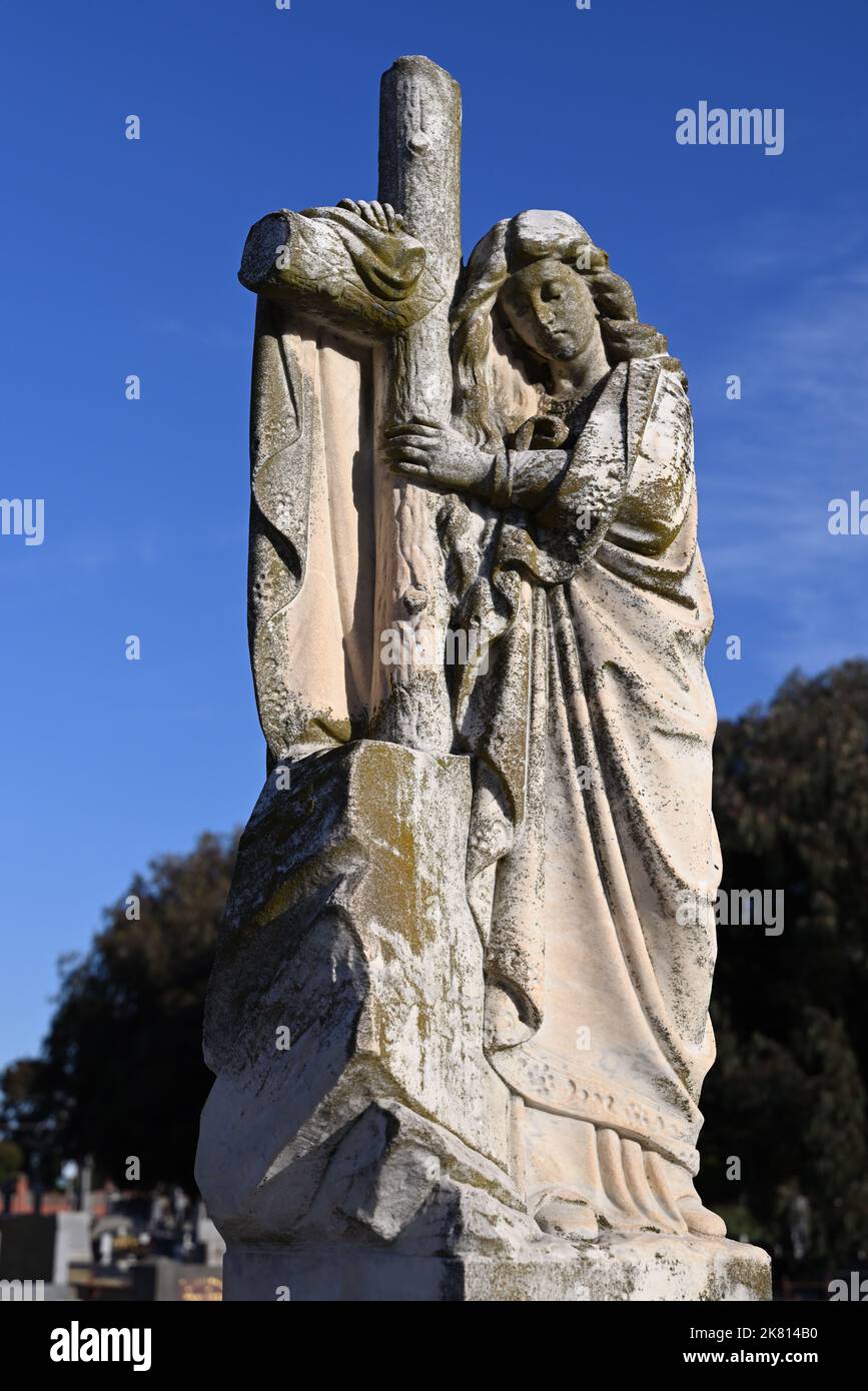 Abgenutzte und moosige Steinskulptur einer Frau, die eine Robe trug, während sie sich auf einem Kruzifix auf einem Friedhof stützte, mit hellblauem Himmel im Hintergrund Stockfoto
