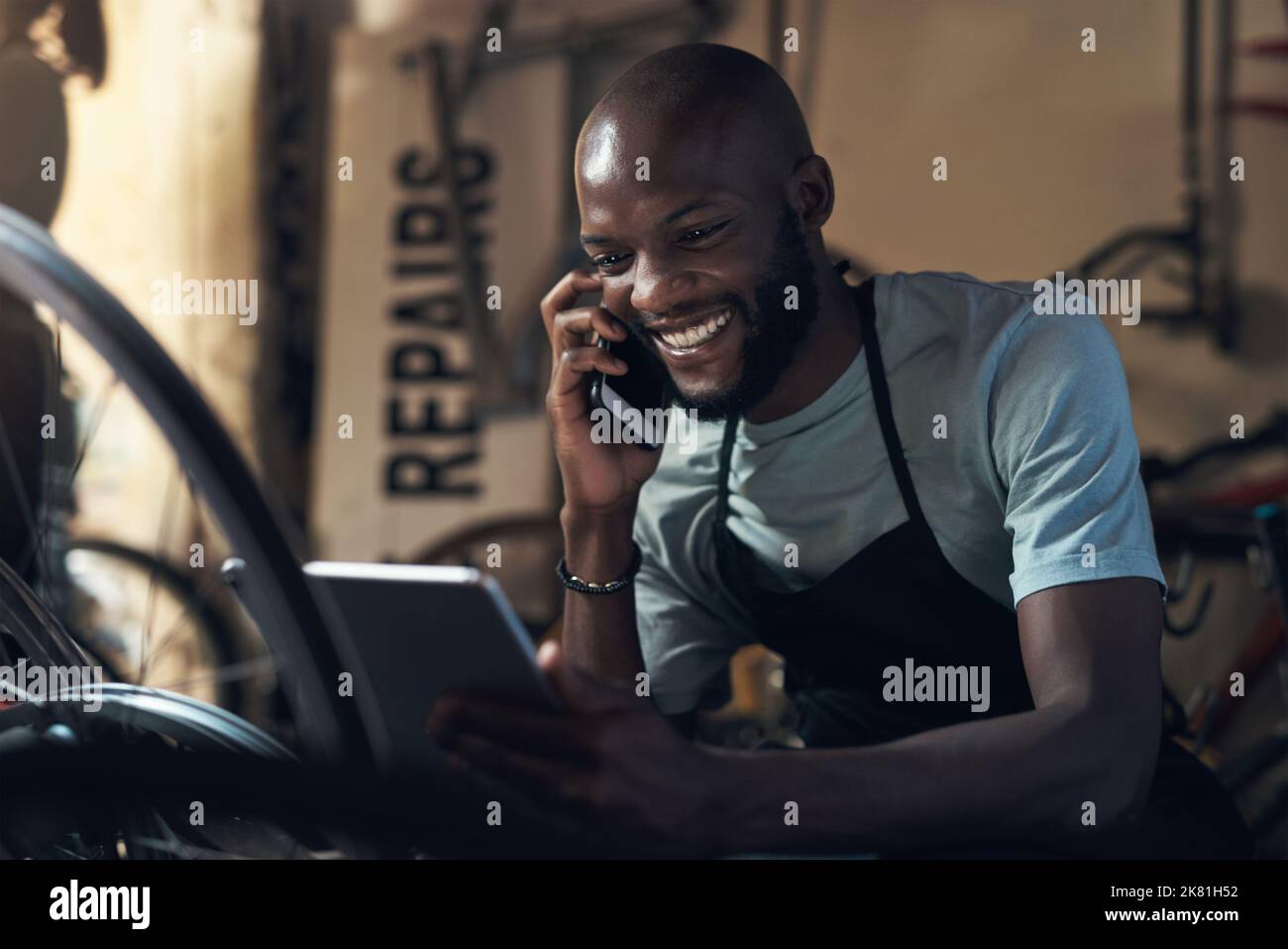 Ich kann nicht glauben, dass ich das verpasst habe. Ein junger Mann, der auf einem Handy sprach und ein digitales Tablet in der Hand hielt, während er in einer Fahrradwerkstatt arbeitete. Stockfoto