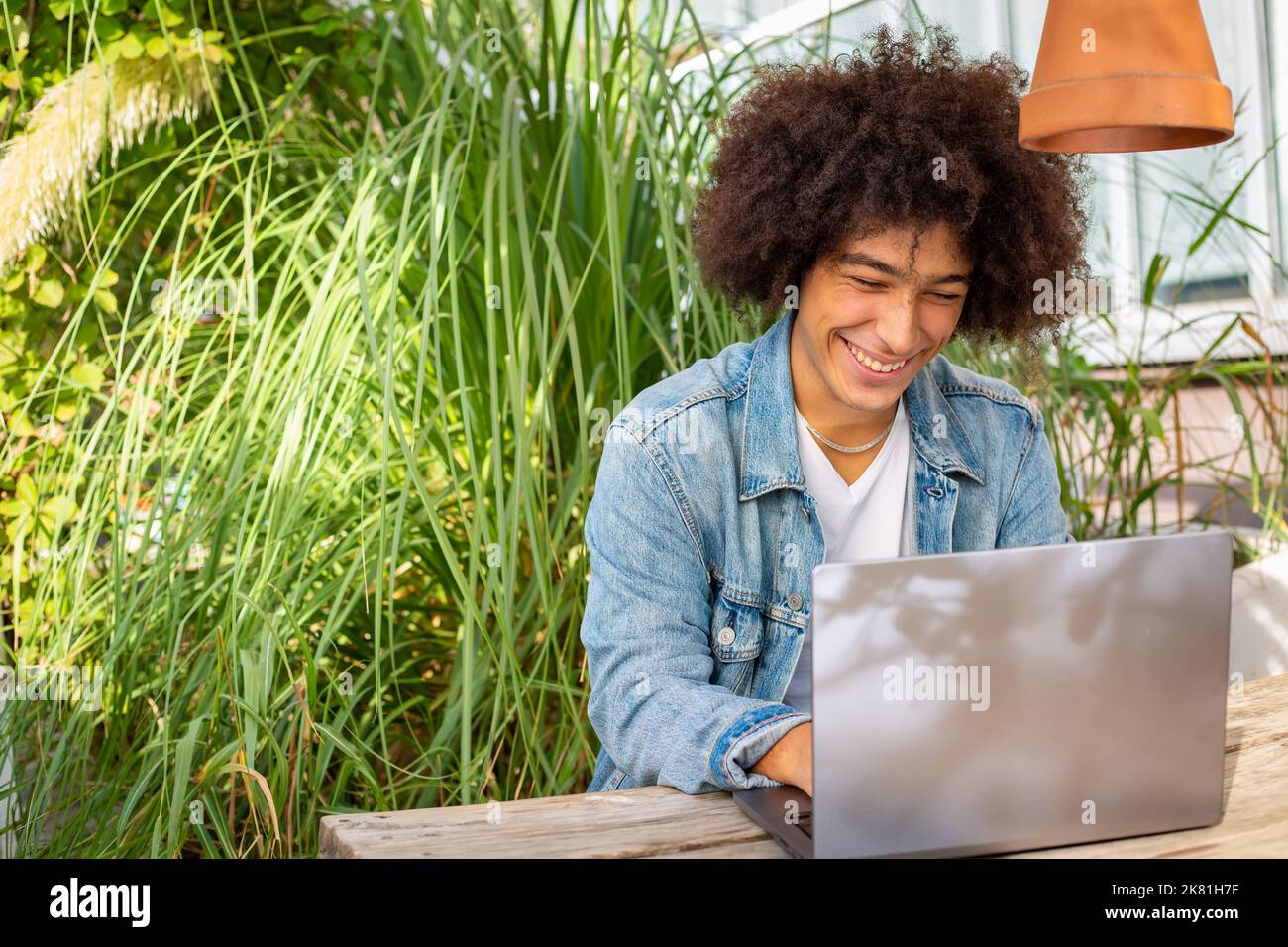 Lächelnder junger ethnischer Typ, 20 Jahre alt, in legerer Kleidung gekleidet, arbeitet auf einem Laptop im Freien, in einer grünen Gegend in der Natur. Grüner Freiraum für Stockfoto