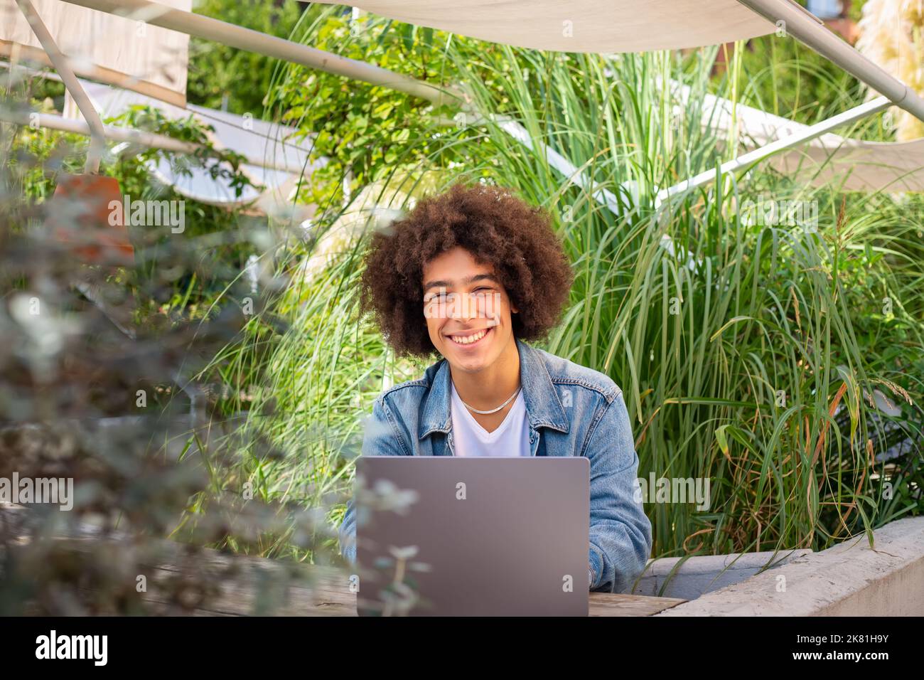 Lächelnder junger ethnischer Typ, 20 Jahre alt, in legerer Kleidung gekleidet, arbeitet auf einem Laptop im Freien, in einer grünen Gegend in der Natur. Grüner Freiraum für Stockfoto