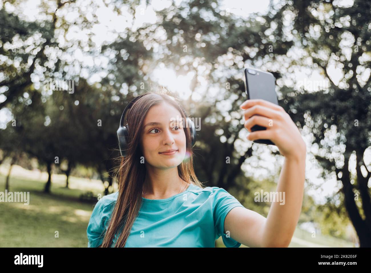 Lächelndes Mädchen im Teenageralter, das ein türkisfarbenes T-Shirt und Kopfhörer trägt und ein Selfie mit ihrem Handy in einem natürlichen Raum macht Stockfoto