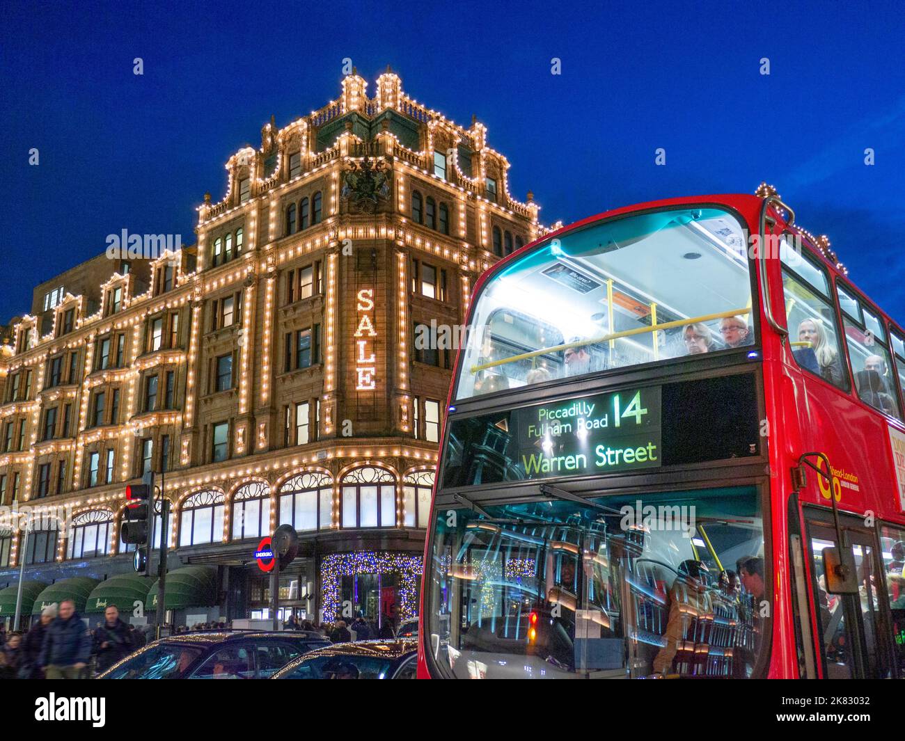 VERKAUF BUS TRANSPORT LONDON SHOPPING Harrods Kaufhaus in der Winternacht Abenddämmerung mit Sale Lights Shopper vorbei an Taxis und roten Bus im Vordergrund auf der Route 14 Knightsbridge London SW1 Stockfoto