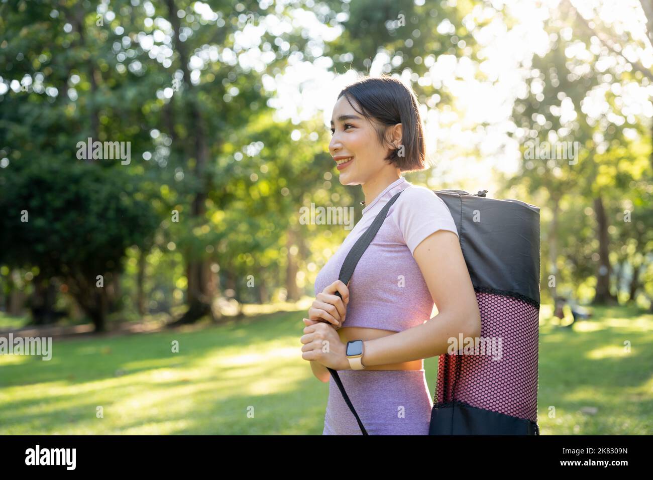 Lächelnde Fitness-Frau, die im Gartenpark steht und eine Yogamatte trägt. Stockfoto