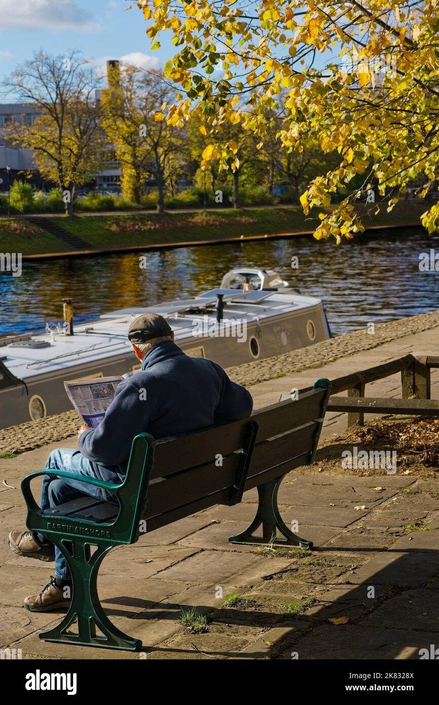 Ein erwachsener Mann, der sich beim Lesen einer Zeitung am Fluss Ouse am Dame Judi Dench Walk, York, North Yorkshire, Großbritannien, entspannt. Stockfoto