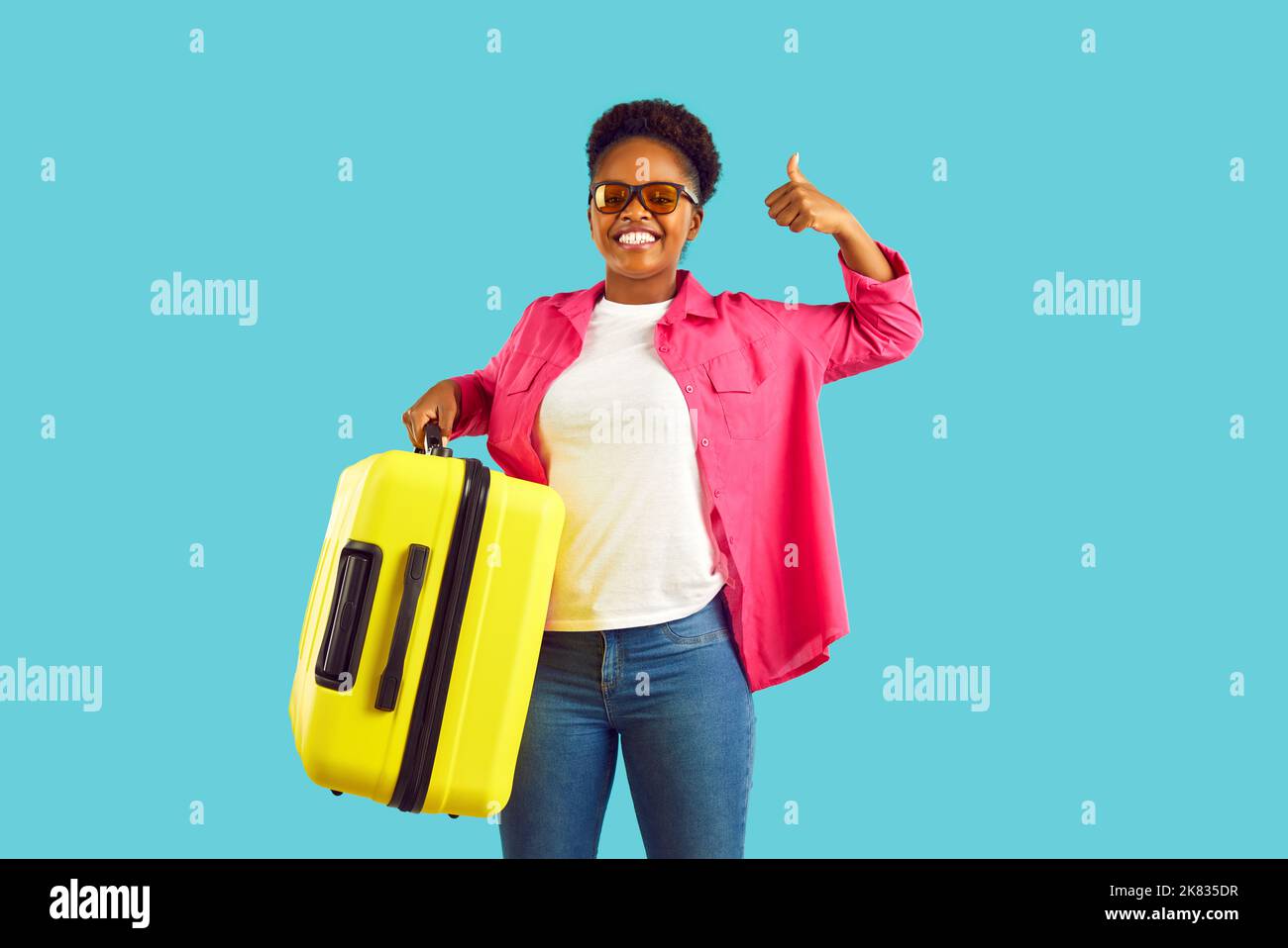 Eine afroamerikanische Frau in einer Sonnenbrille zeigt den Daumen mit einem gelben Koffer auf türkisfarbenem Hintergrund. Stockfoto Eine afroamerikanische Frau in einer Sonnenbrille zeigt den Daumen mit einem gelben Koffer auf türkisfarbenem Hintergrund. Stockfoto