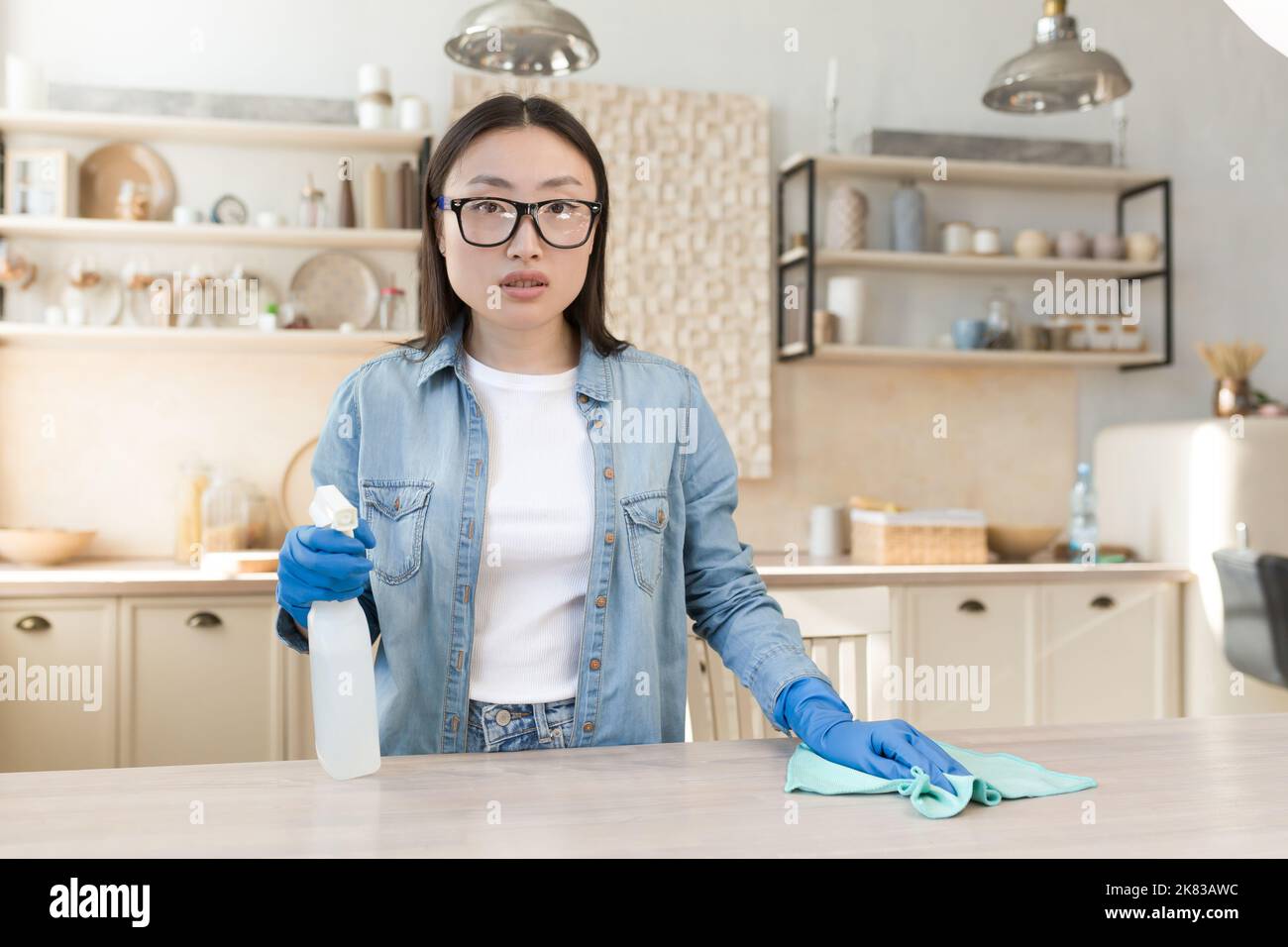 Portrait einer jungen asiatischen Hausfrau, die zu Hause putzt. Wischt den Tisch in der Küche mit einem Lappen in Gummihandschuhen, verwendet Haushaltschemikalien. Er steht, hält ein Spray und schaut auf die Kamera. Stockfoto
