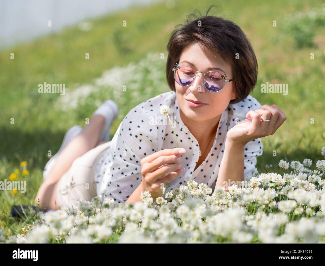 Frau in farbenfroher Sonnenbrille schnuppert Kleeblüten auf dem Rasen im Stadtpark. Natur in der Stadt. Entspannen Sie sich nach der Arbeit im Freien. Sommerliche Stimmung. Stockfoto