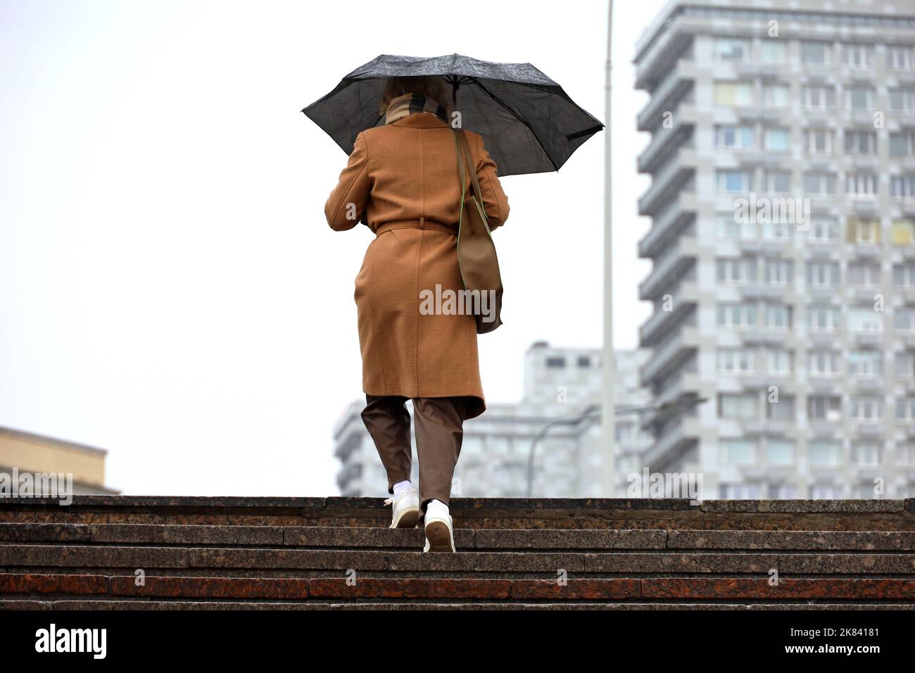 Frau mit Regenschirm, die auf dem Hintergrund der Gebäude die Stufen hinauf läuft. Regen in der Herbststadt Stockfoto