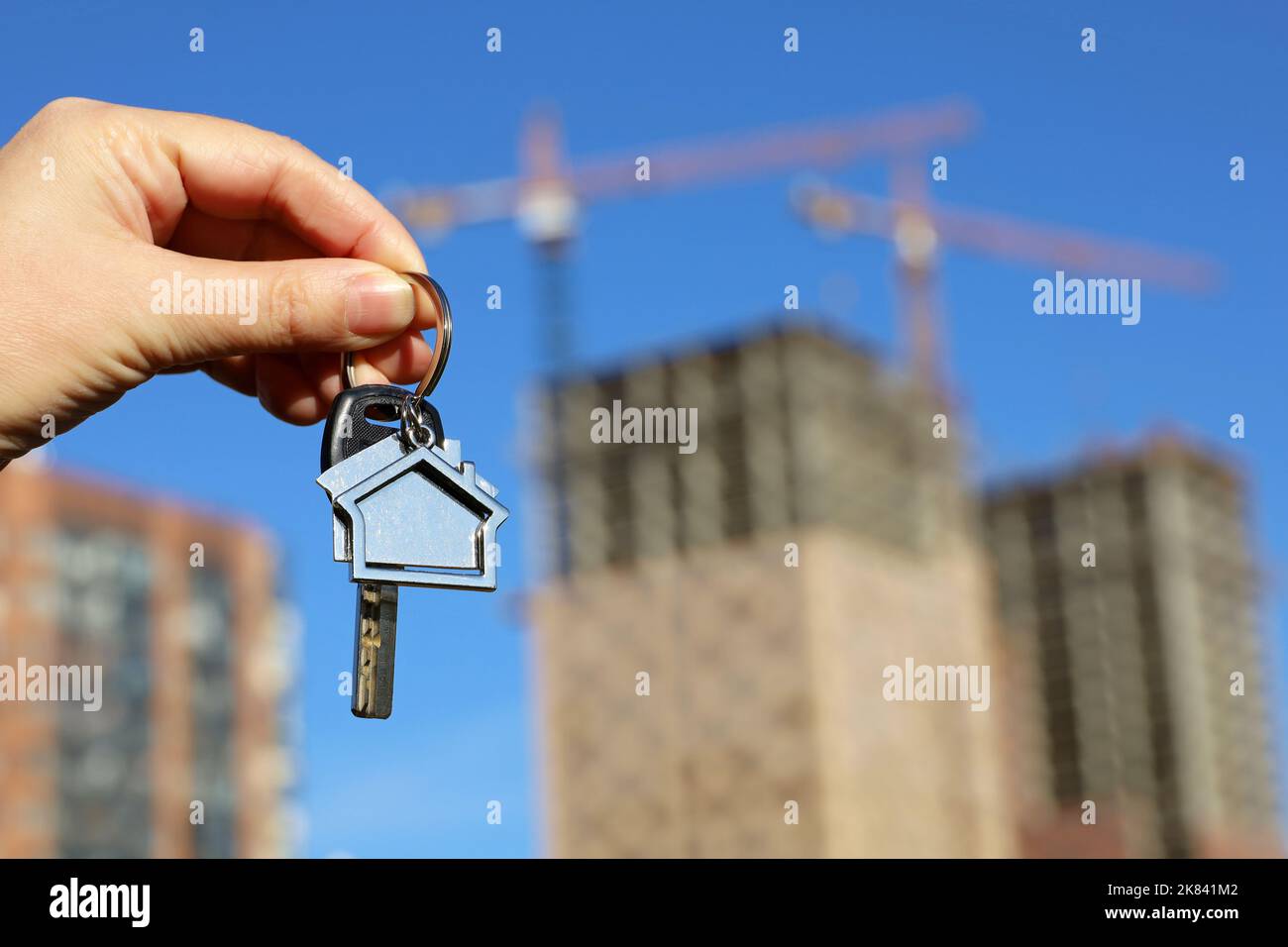 Immobilienmakler, Schlüsselanhänger in Form eines Hauses und Schlüssel in weiblicher Hand auf dem Hintergrund von Baukräne und Neubauten. Byung Wohnung oder Miete Stockfoto