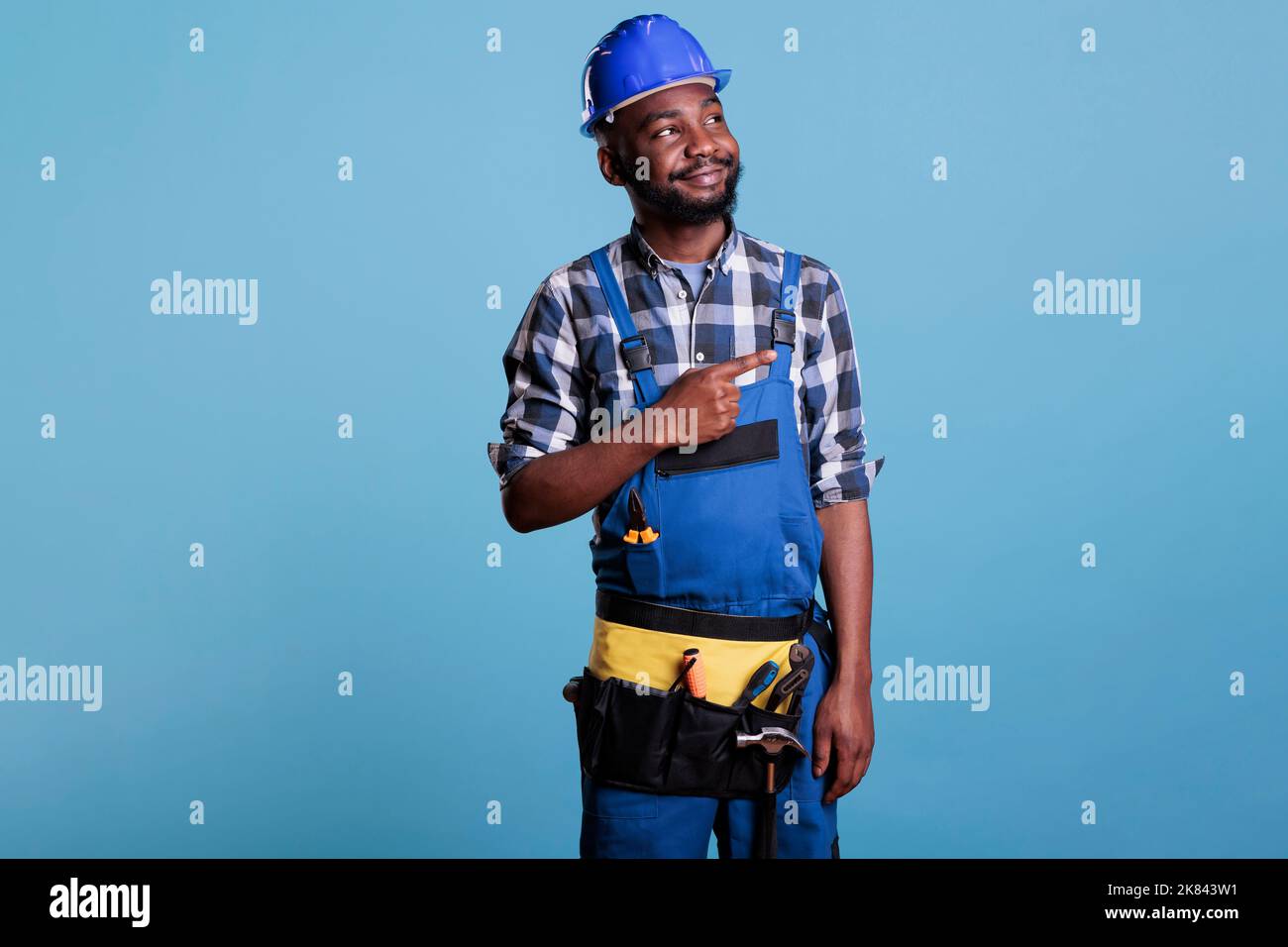Porträt eines glücklichen Bauarbeiters bei der Arbeit, der einen harten Hut trägt und seinen Finger zur Seite zeigt. Elektriker trägt Gürtel mit Werkzeugen im Studio vor blauem Hintergrund aufgenommen. Stockfoto