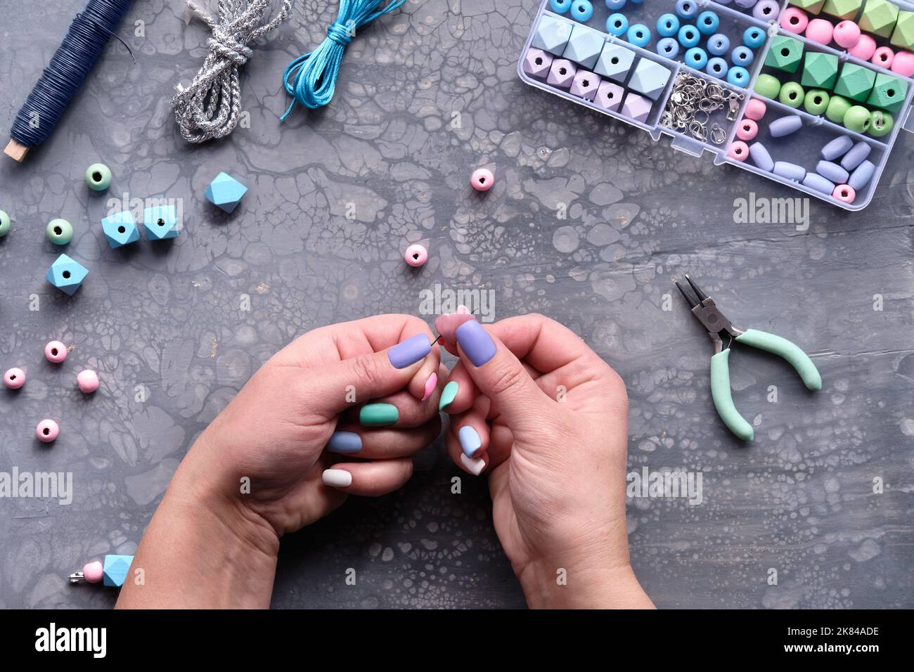 Handgefertigter Schmuck. Schachtel mit Perlen, Hände mit Werkzeug. Machen handgemachten Schmuck für sich selbst oder als selbst gemachte benutzerdefinierte Geschenke. Flach auf grauem Strukturmuster Stockfoto