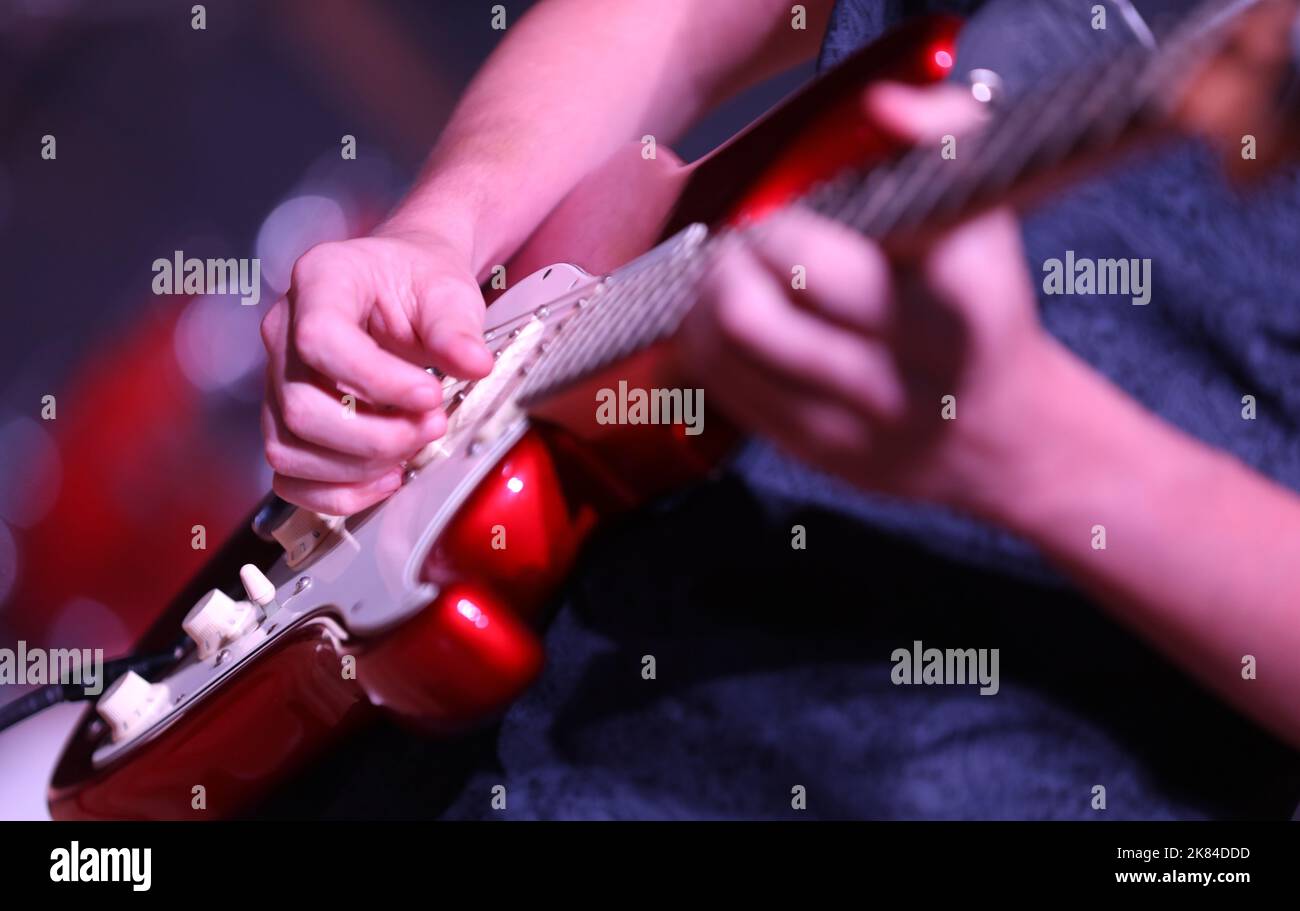 Nahaufnahme von Hand und Fingern, die Musik auf einer leuchtend roten E-Gitarre spielen. Hand-Geklimper ärgern sich mit einem absichtlich verschwommenen Spotlight-Hintergrund Stockfoto