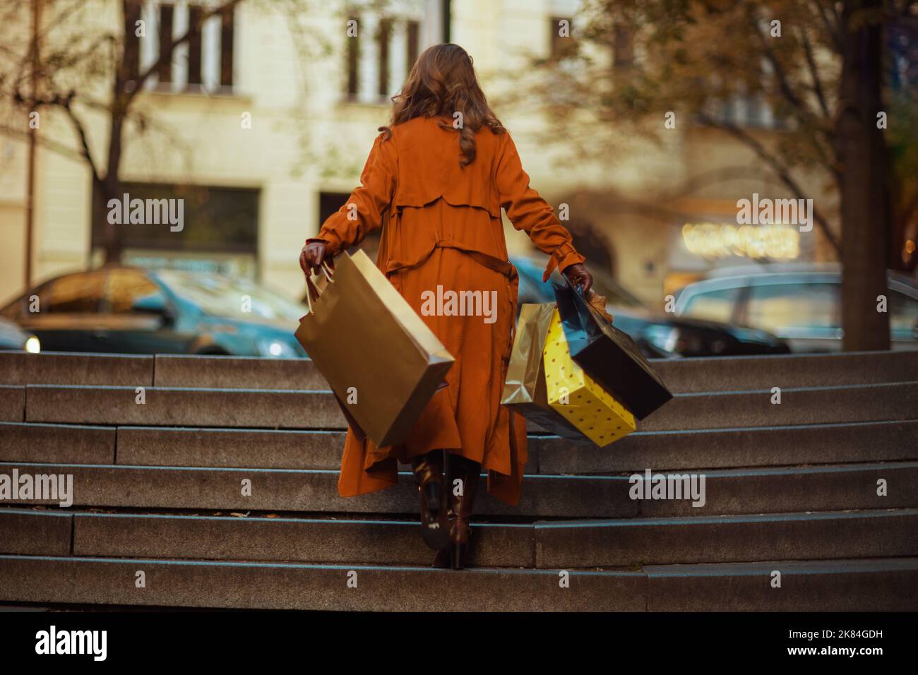 Hallo oktober. Von hinten gesehen junges Weibchen in orangefarbenem Trenchcoat mit Einkaufstaschen in der Stadt. Stockfoto