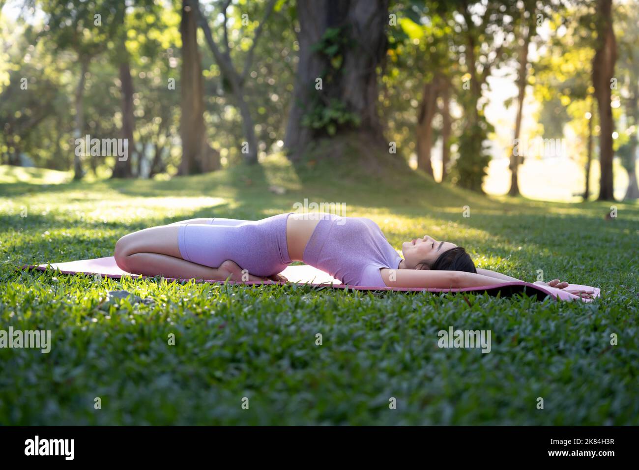 Attraktive junge asiatische Frau praktiziert Yoga, trainiert im Park, steht ein Bein auf einer Yogamatte und zeigt eine ausgeglichene Haltung. Wellness-Lifestyle und Stockfoto
