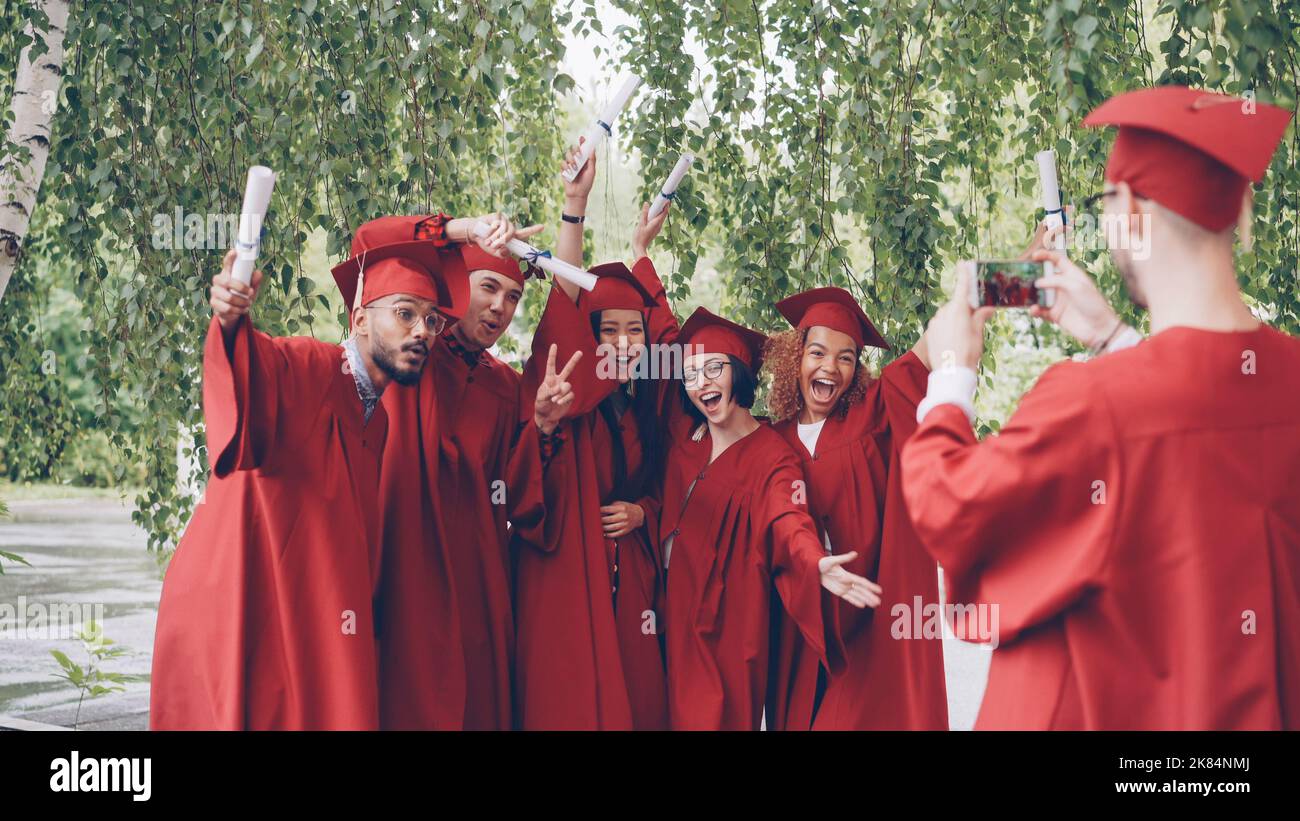 Der graduierende Student nimmt ein Video seiner Freunde in Kleidern und Leichenbrettern auf, die Diplome halten, die Hände winken und posieren und auf die Smartphone-Kamera blicken. Menschen- und Technologiekonzept. Stockfoto