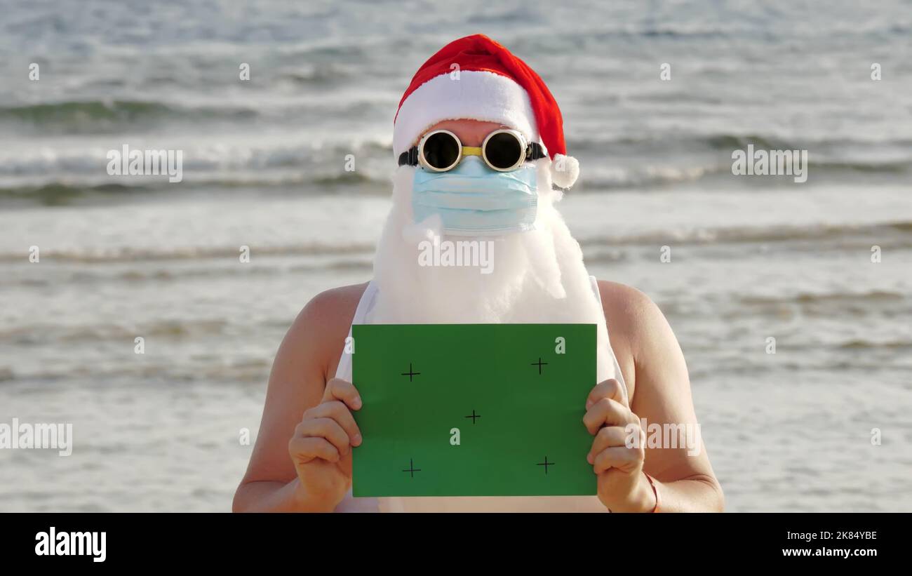 Witziger weihnachtsmann, in einer Schutzmaske. Santa, in Sonnenbrille, mit einem grünen Schild, vor dem Hintergrund des Meeres. santa claus Urlaub während Coronavirus Pandemie oder Lockdown, an der Küste. Hochwertige Fotos Stockfoto