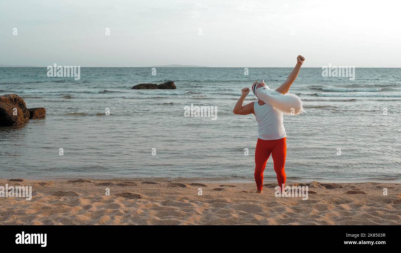 Lustiger weihnachtsmann. weihnachtsmann am Meer, im Sommerurlaub. weihnachtsmann tanzt, springt am Strand, bei Sonnenaufgang. weihnachtsmann in einem T-Shirt und roten Leggings am Meer. Hochwertige Fotos Stockfoto