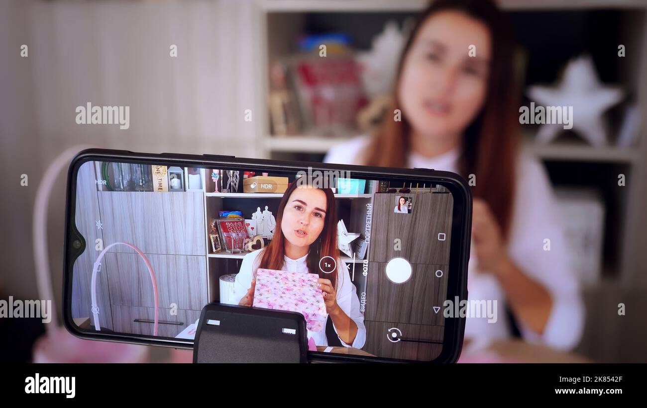 Geschenkverpackung. Attraktive Frau, Designer von Geschenken und Dekorationen zeigt Training online, zeichnet Videoblog, Webinar, sitzt vor dem Smartphone, Kamera. Online-Verkauf. Hochwertige Fotos Stockfoto