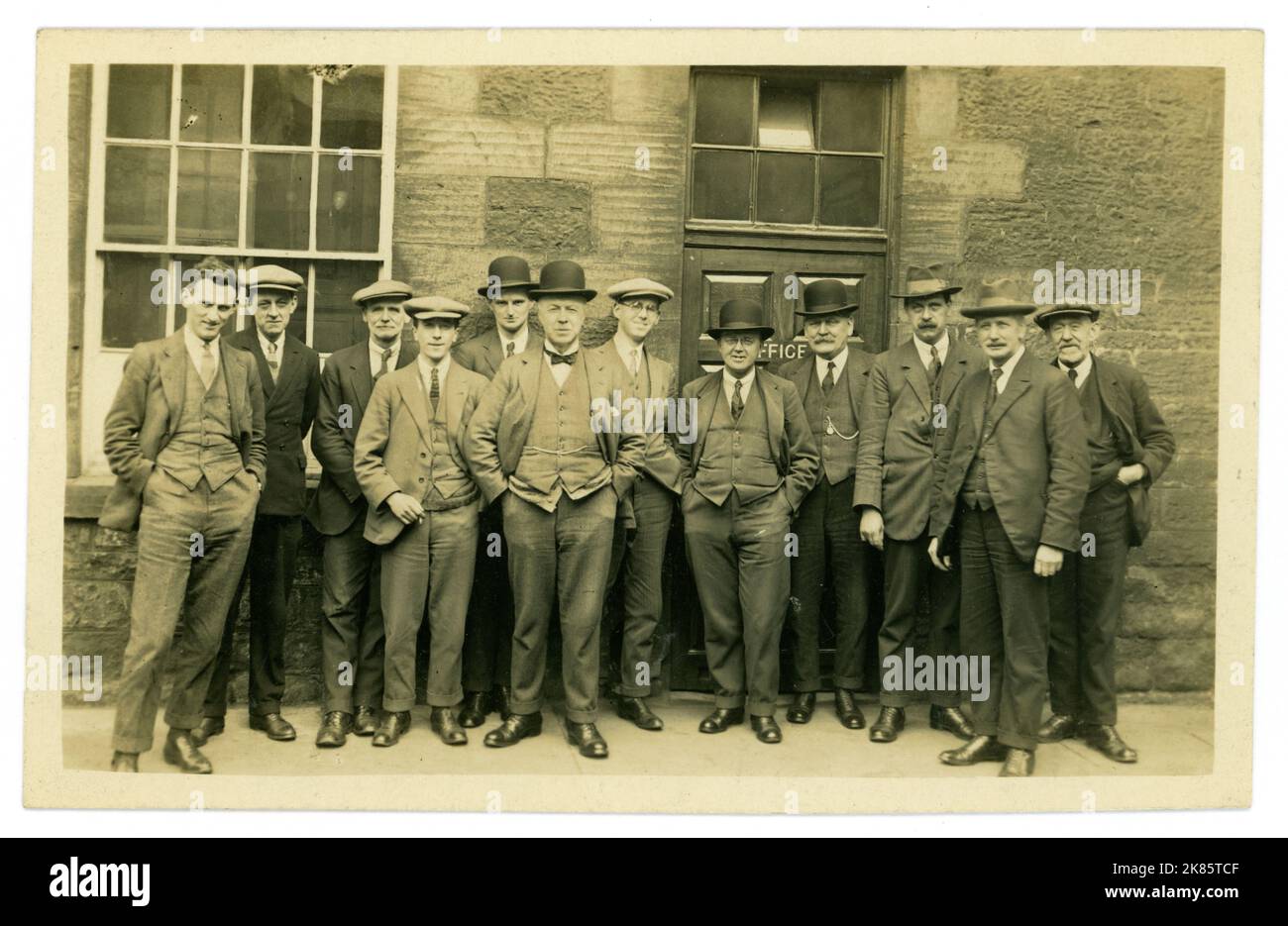 Originalpostkarte aus den 20er Jahren, stellenweise getönte Sepia, Büroangestellte, eine Gruppe von Männern der Arbeiterklasse, die vor ihrem Büro stehen, viele Charaktere, die Anzüge und verschiedene Hüte tragen - flache Kappen, Melone und Homburger Hüte. Kurz vor Beginn der Großen Depression im Jahr 1929. Vom 1928. April, Großbritannien Stockfoto