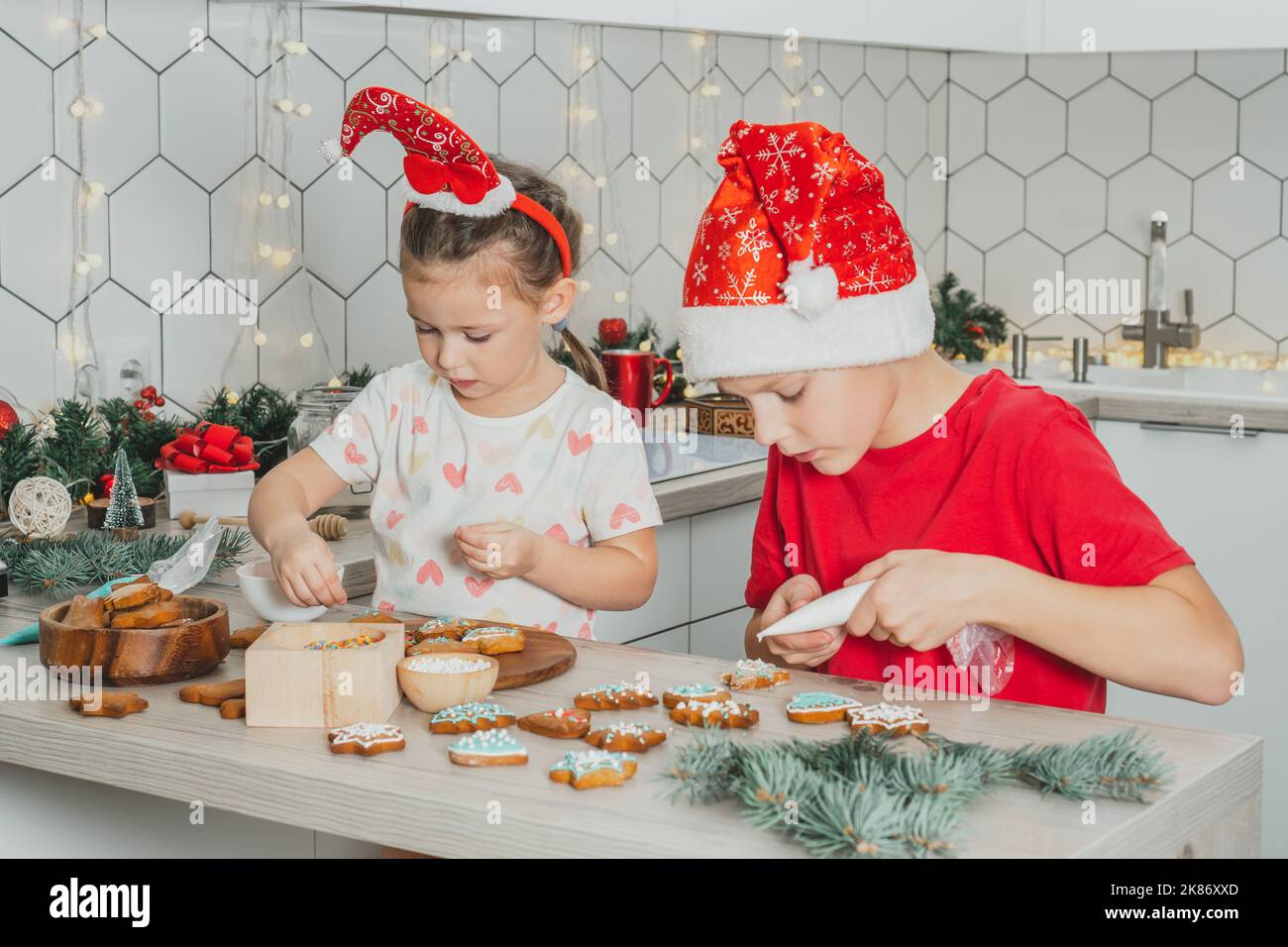 Kleine dunkelhaarige Mädchen 3 Jahre alt und Jungen 8 Jahre alt in roten Santa Hut schmücken Lebkuchen mit Glasur. Geschwister in weißen Weihnachten Stockfoto