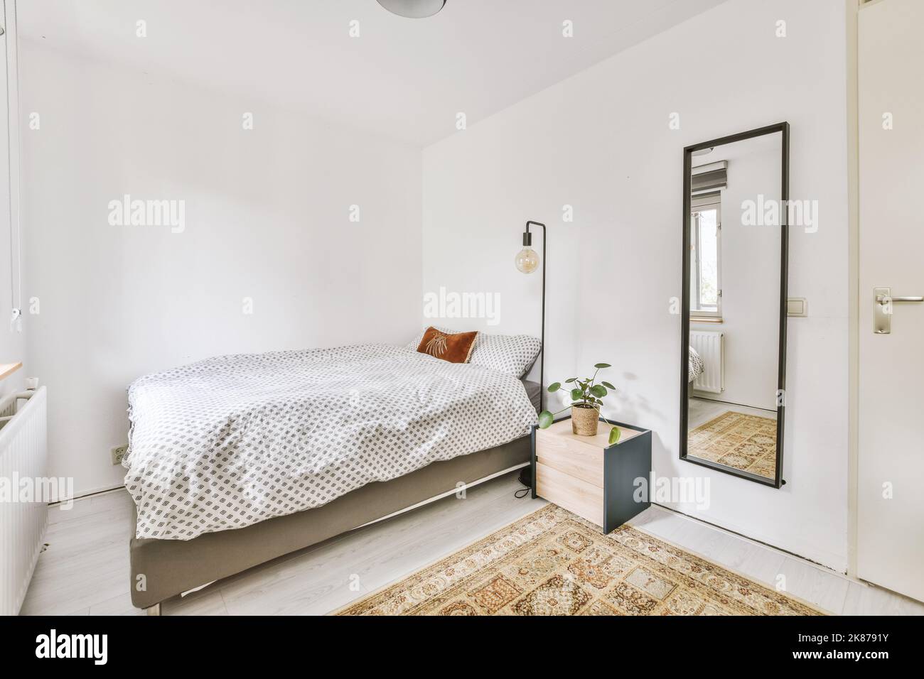 Komfortables Bett und Lampe in einem kleinen, schmalen Schlafzimmer im minimalistischen Stil mit weißen Wänden und schwarzen Vorhängen am Fenster in einem modernen Apartment Stockfoto