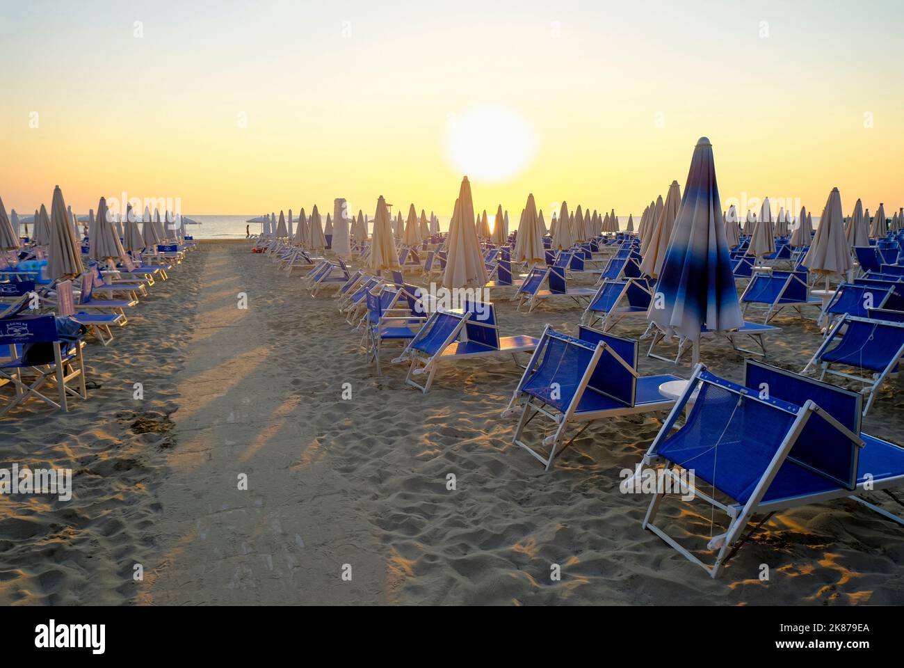 Sonnenschirme und Sonnenliegen am Strand über dem Sonnenaufgangshimmel. Weg auf dem Sand zum Strand. Sommerurlaub Hintergrund. Senigallia, Italien Stockfoto