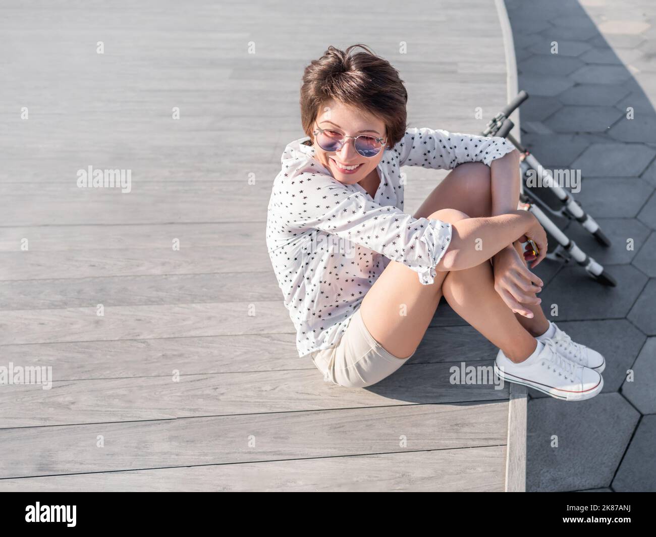 Wind kräuselt kurze Haare der Frau in bunten Sonnenbrillen. Lächelnde Frau bei offener Holzszene. Sommerliche Stimmung. Aufrichtige Emotionen. Kick Scooter - Öko-Freund Stockfoto