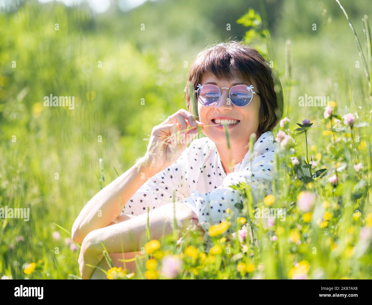 Frau in bunten Sonnenbrillen, genießt Sonnenlicht und Blumenduft auf Grasfeld. Sommerliche Stimmung. Entspannen Sie sich im Freien. Selbstberuhigend. Stockfoto