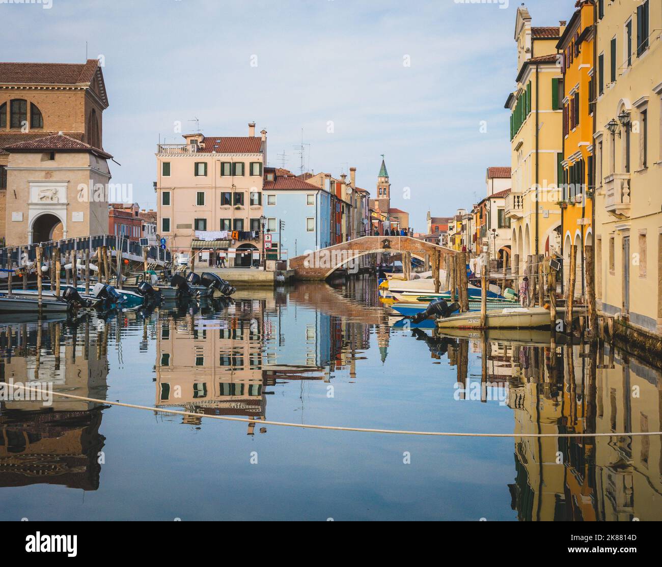 Chioggia Stadtbild mit schmalen Wasserkanal mit festgetäuten Booten, Gebäuden, Backsteinbrücke und Turm der Kirche San Giacomo Apostolo – venezianische Lagune, Italien Stockfoto