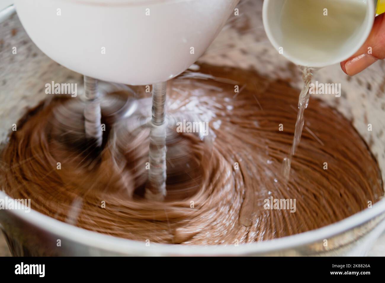 Nahaufnahme einer Tasse Eier, die mit eingeschaltetem Mixer in eine Rührschüssel mit Schokoladenkuchen gedumpt werden. Stockfoto