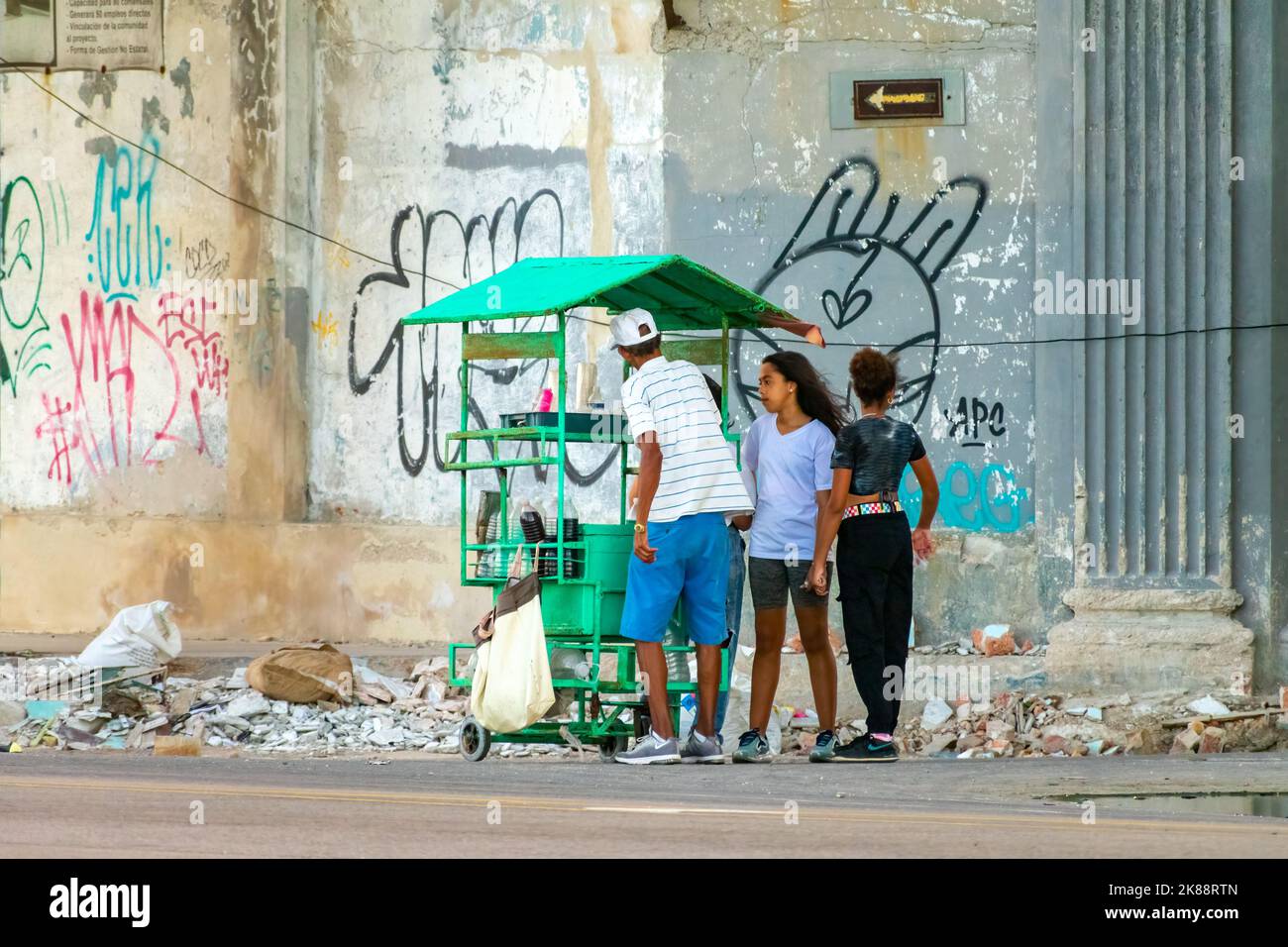 Zwei Frauen kaufen aromatisiertes Eis aus einem kleinen Business-Cart in einer Stadtstraße. In der Wand hinter den Menschen sieht man Graffiti mit städtischer Kennzeichnung Stockfoto