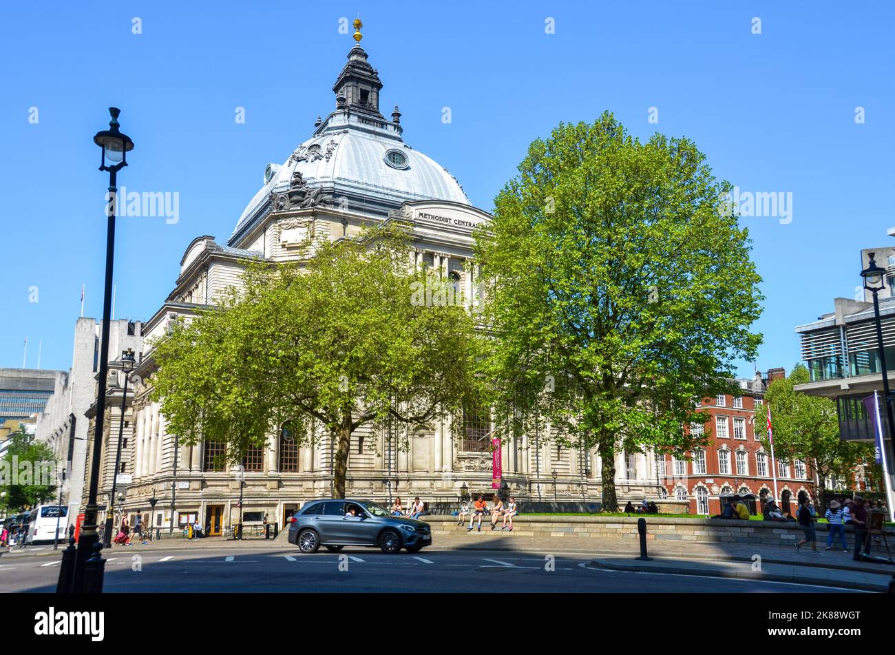 Ein Blick auf die Central Hall Westminster im Hintergrund mit zwei großen Bäumen, Menschen und einem Auto, das in London vorbeifährt. Stockfoto