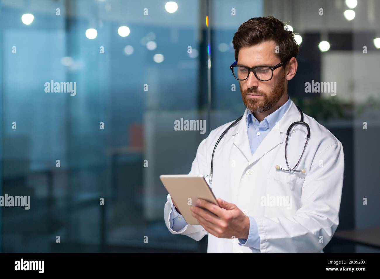 Ein seriöser und nachdenklicher Arzt liest medizinische Informationen von einem Tablet-Computer, ein Mann in einem medizinischen Kleid verwendet eine Tablette, konzentriert sich, gibt Nachrichten ein und überprüft die Patientenakte Stockfoto
