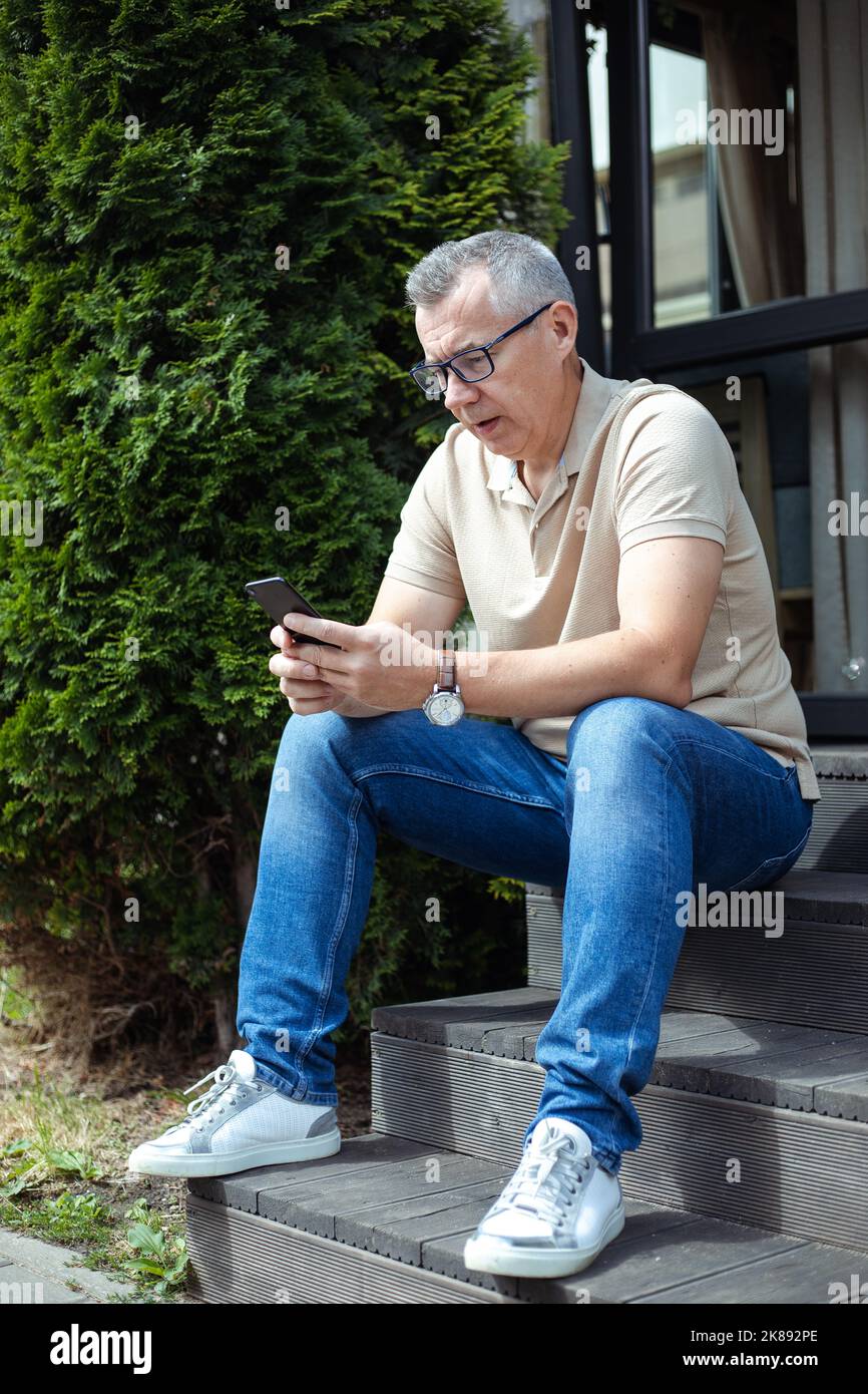 Trauriger, frustrierter älterer Mann, der in einer Brille schielt, benutzt Smartphone und sitzt auf einer Treppe in der Nähe des Eingangs. Technophobe brauchen Hilfe Stockfoto