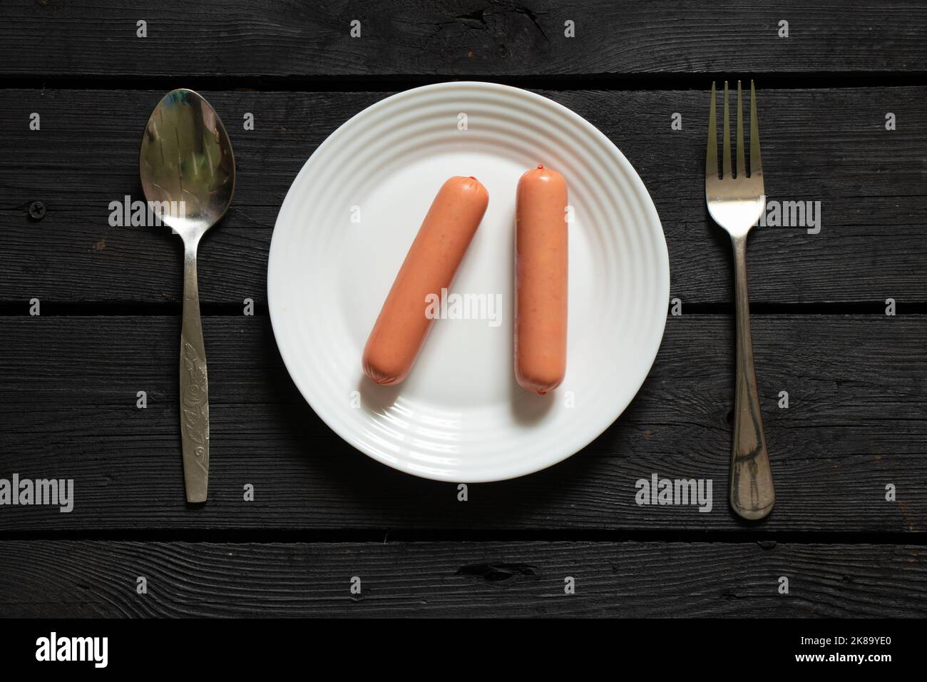 Zwei Rohwürste liegen auf einem weißen Teller neben einem Löffel und eine Gabel auf einem schwarzen Holztisch, Junk Food, Hunger und Armut, Halbzeug Stockfoto