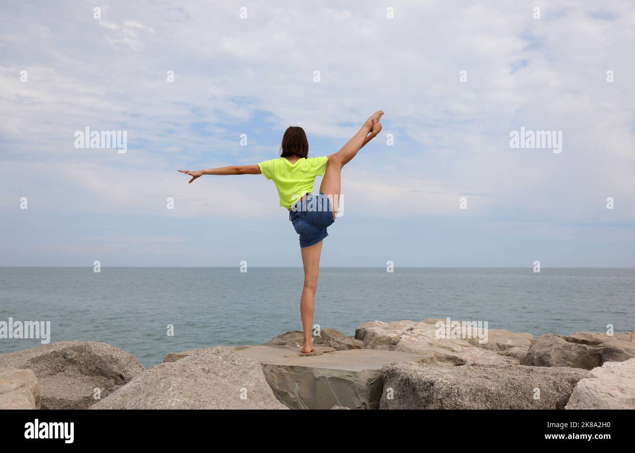 Das junge Mädchen macht Gymnastik, um ihr Gleichgewicht zu trainieren, indem es auf einem Bein auf den Felsen am Meer belastet Stockfoto