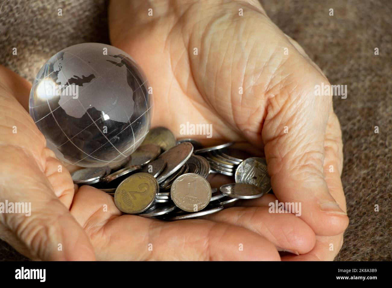 Ukrainische Groschen und eine Glaskugel des Planeten liegen in den Händen einer alten Frau, Finanzwelt und Menschen Stockfoto