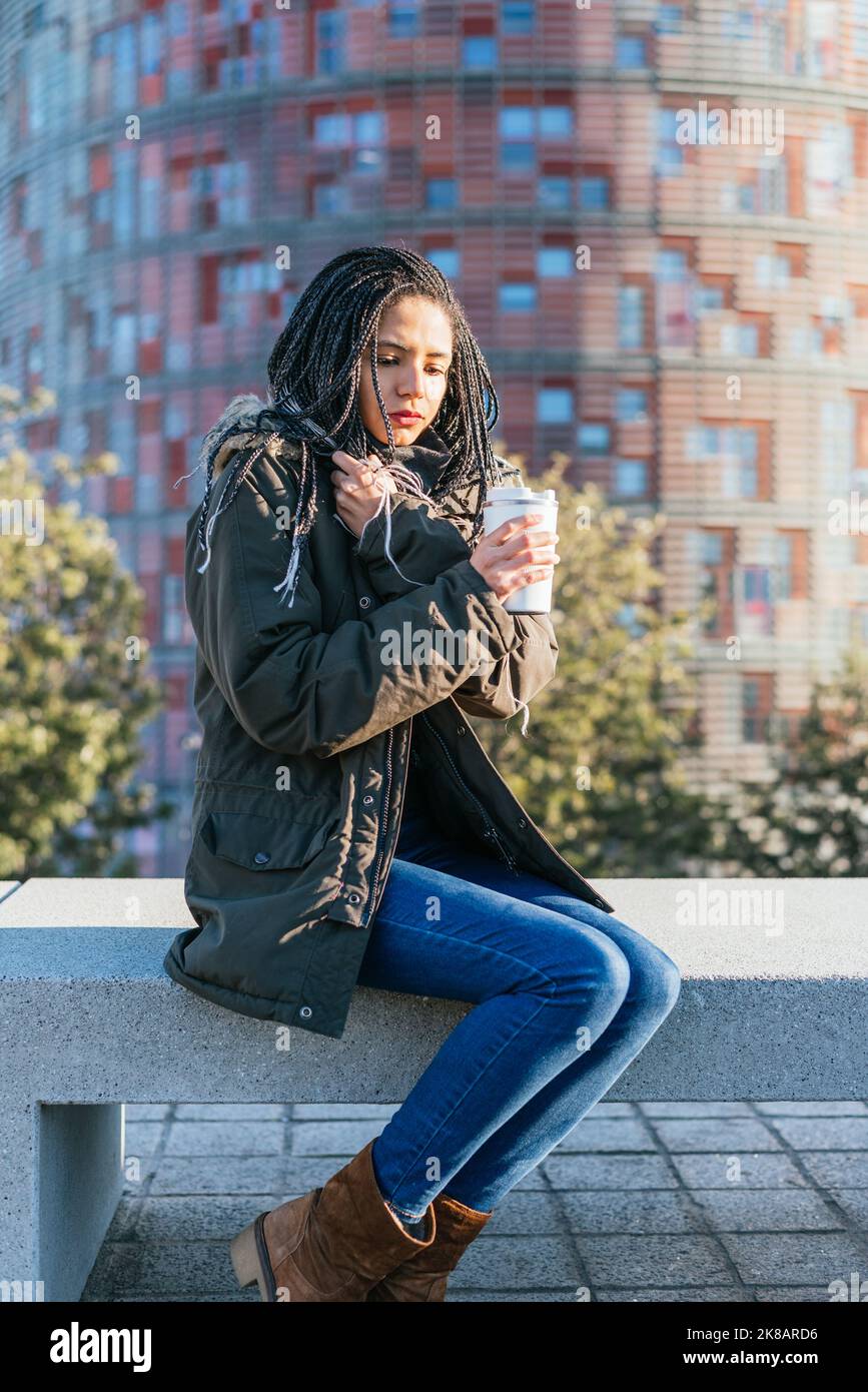 Nachdenkliche hispanische Hündin in warmer Oberbekleidung mit Dreadlocks, die auf einer Steinbank mit Thermoskanne mit heißem Getränk in der Stadt sitzen Stockfoto
