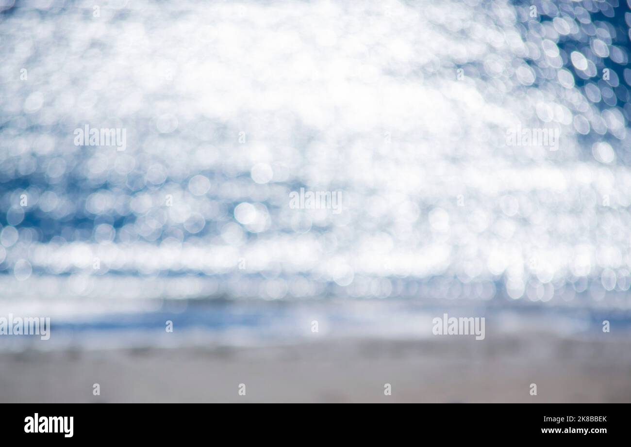 Unscharfer Hintergrund. Meereswelle mit weißem Schaum am Sandstrand am sonnigen Morgen des Sommers. Natürliche, unscharfe Bokeh-Textur im Hintergrund. Schöner natürlicher Hintergrund. Umwelt, globale Erwärmung. Stockfoto