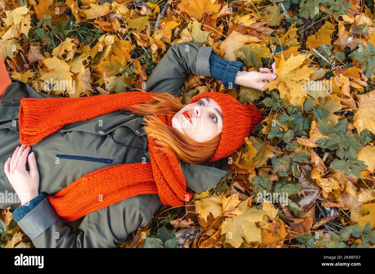 Verträumte Frau mit herbstlichen gelben Blättern in der Hand in orangefarbenem Strickhut und auf Blättern im Park liegendem Scarfl Stockfoto