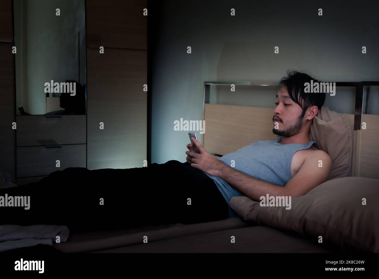 Asiatischer junger Mann mit Mobiltelefon auf dem Bett in der Nacht. Stockfoto