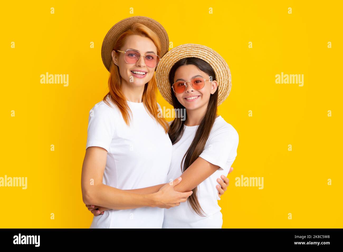 Fröhliche, stilvolle Mutter und Tochter posieren auf gelbem Hintergrund im Studio, tragen Strohhut und Sonnenbrille. Porträt der Familie im Sommer. Stockfoto