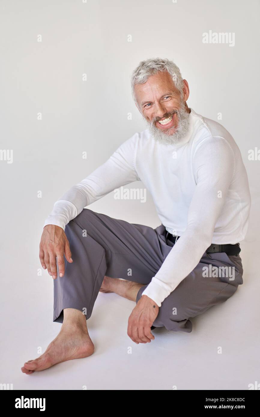Lächelnder, bärtiger älterer Mann, der auf dem Boden an der weißen Wand sitzt. Stockfoto