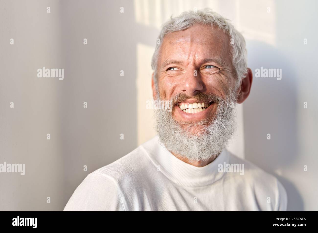 Lächelnder bärtiger älterer Mann, der lachend an der weißen Wand im Sonnenlicht steht. Stockfoto