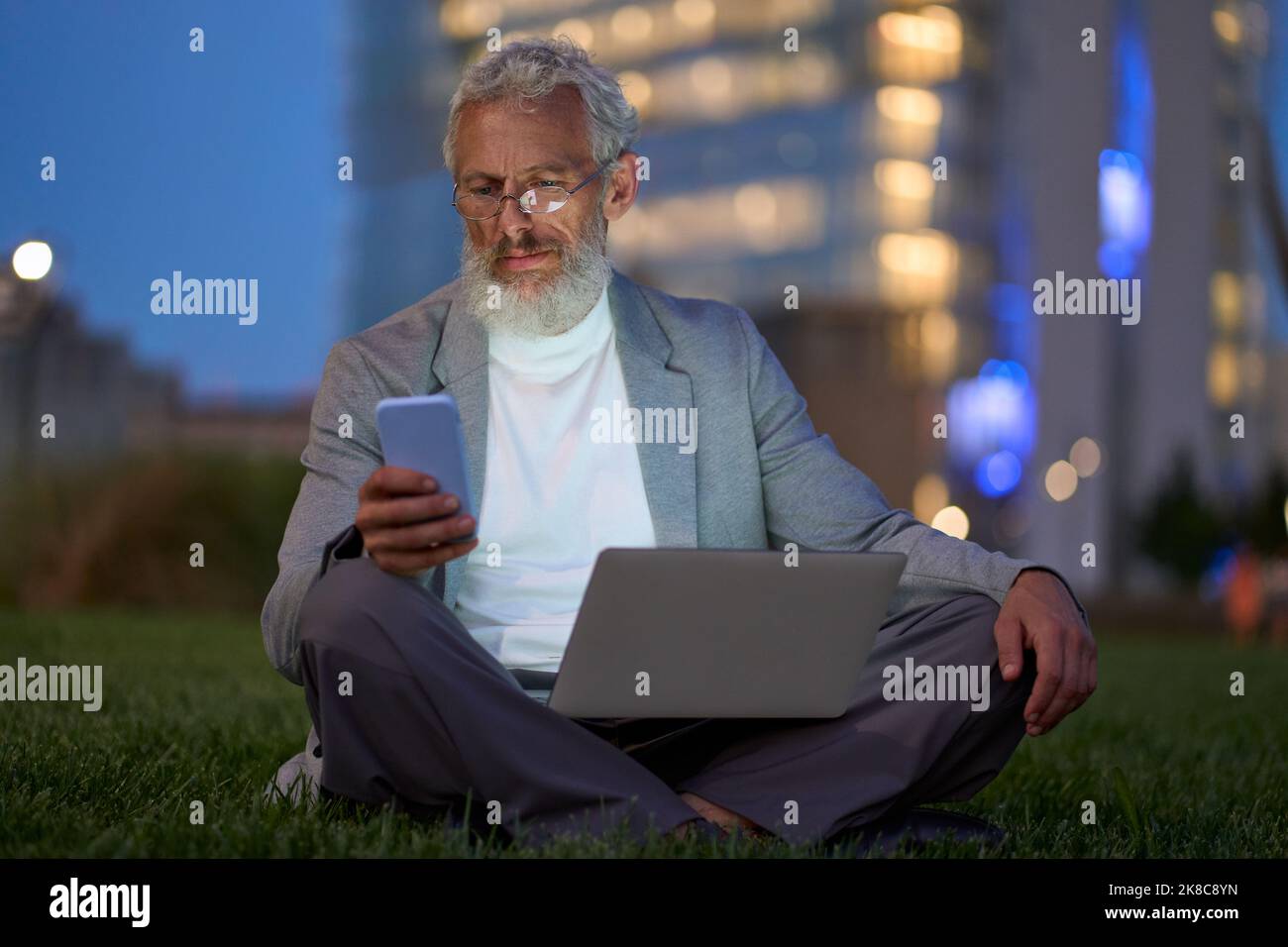 Alter Geschäftsmann, der nachts mit dem Smartphone vor dem Büro im Stadtpark sitzt. Stockfoto