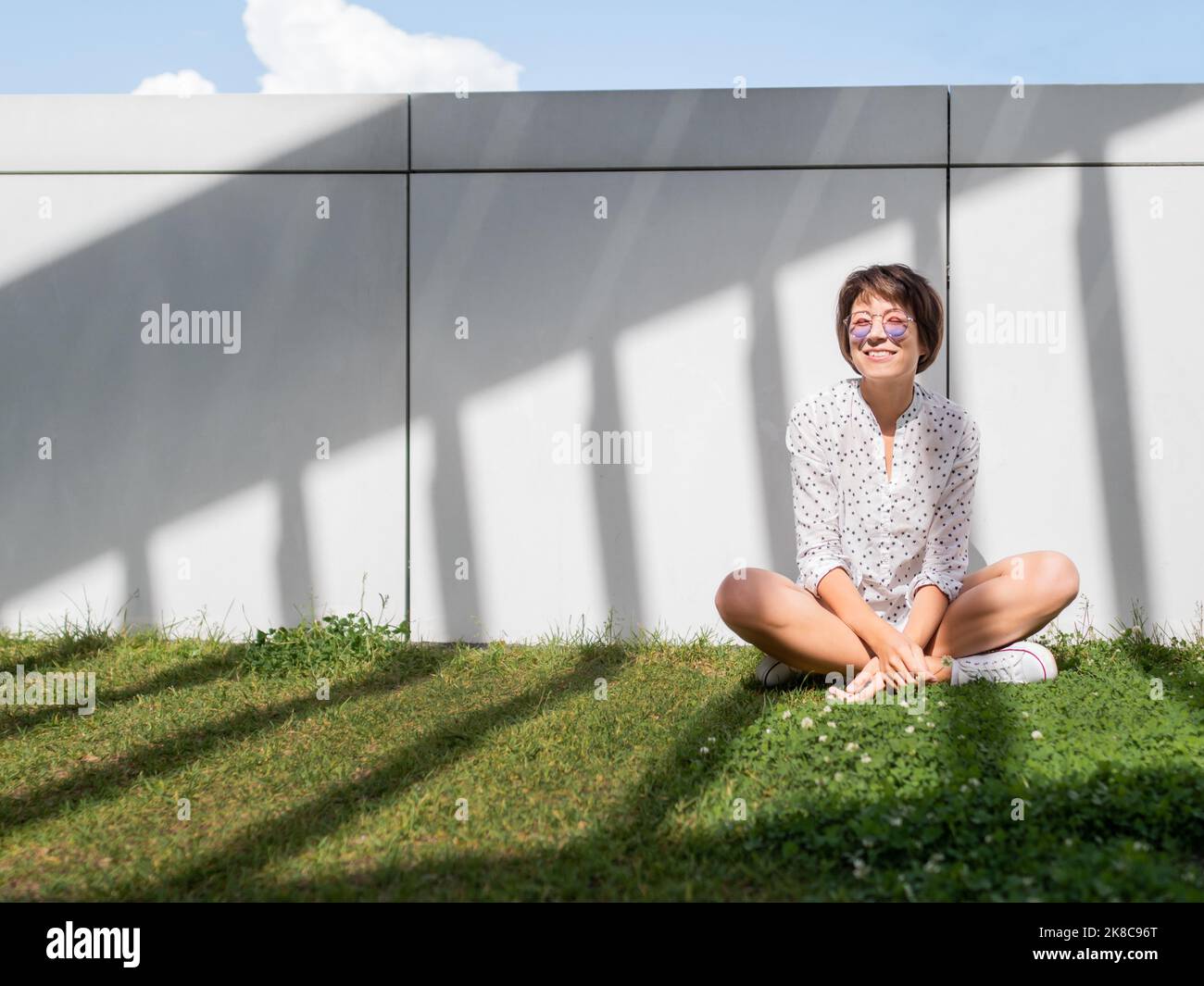 Lächelnde Frau in farbenfroher Sonnenbrille ruht sich auf dem Rasen im Stadtpark aus. Natur in der Stadt. Entspannen Sie sich nach der Arbeit im Freien. Sommerliche Stimmung. Stockfoto