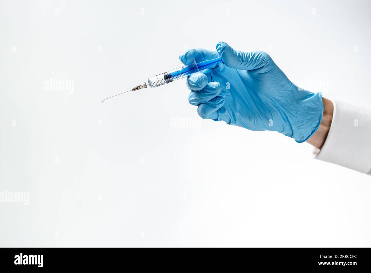 Spritze mit einem Tropfen auf einer Nadel in der Hand in einem blauen Gummihandschuh auf weißem Hintergrund. Medizinisches Injektionskonzept. Stockfoto