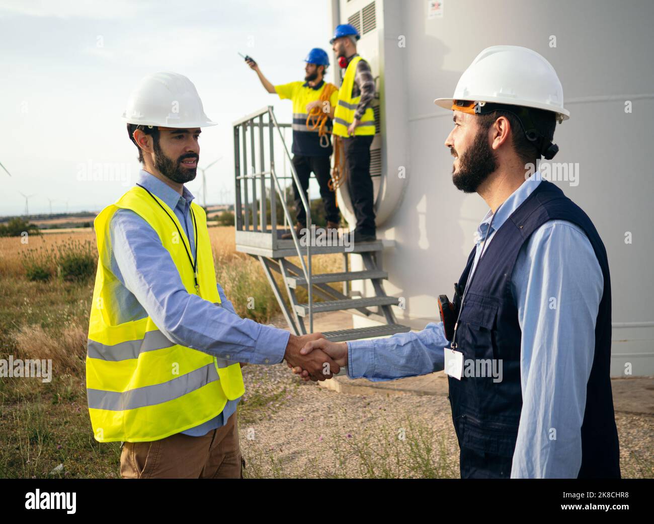 Zwei männliche Ingenieure schütteln sich auf einem Feld von Windmühlen die Hände. Abkommen, erneuerbare Energien, Energiewende Stockfoto